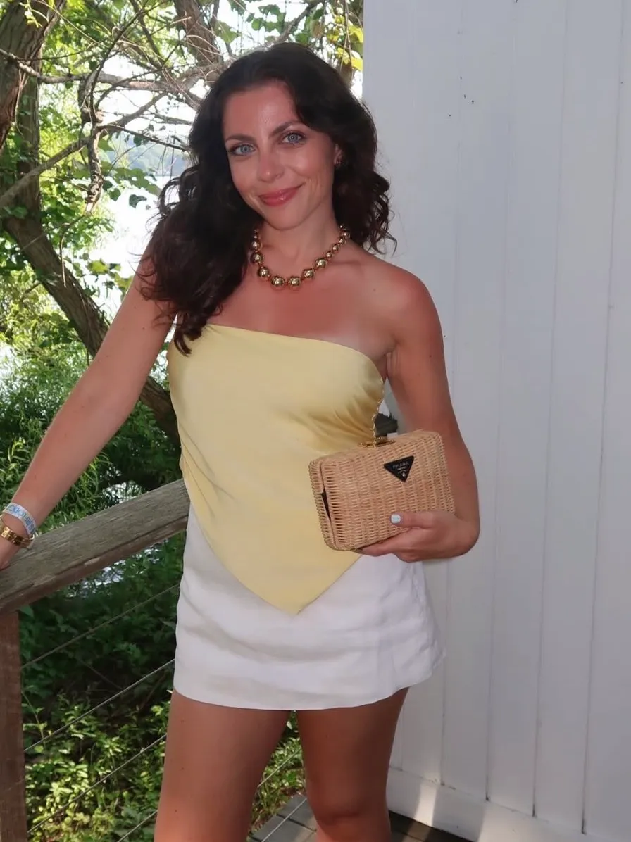 A woman smiling while wearing a yellow strapless top, white skirt, and holding a Prada straw handbag.