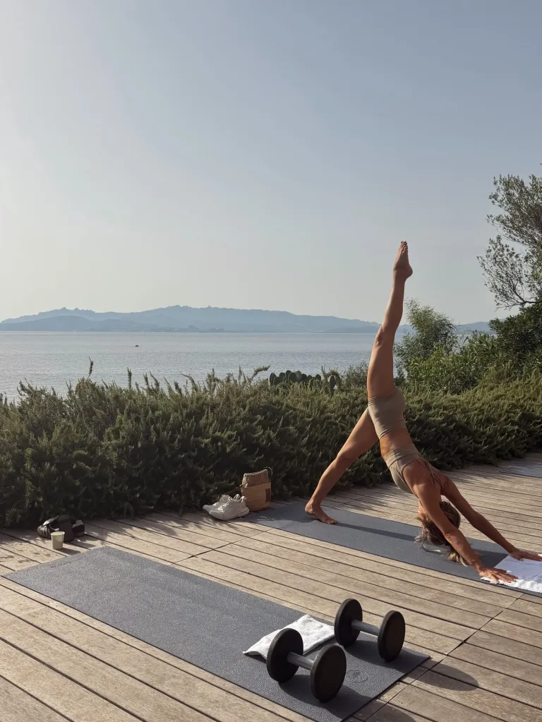 Julianne Hough in a three-legged downward-facing dog yoga pose on a wooden deck overlooking the ocean.