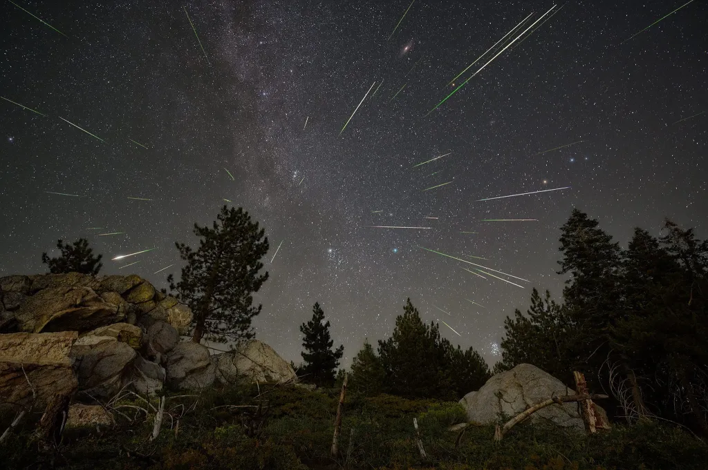 The Milky Way and numerous bright meteors streak across a dark night sky over a rocky, forested landscape.