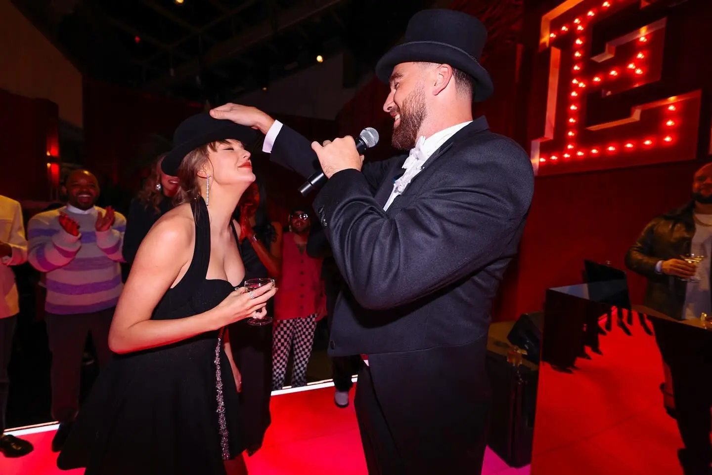Travis Kelce in a top hat putting a fedora on Taylor Swift's head.