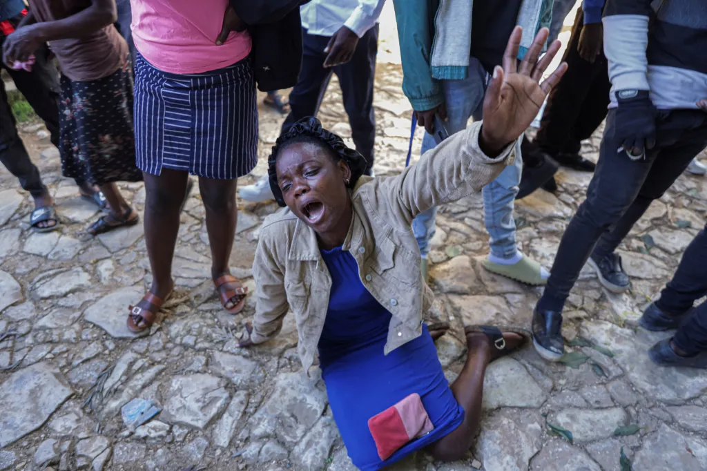 A woman kneeling on a cobblestone road, distraught and screaming with her hand raised, surrounded by other people.