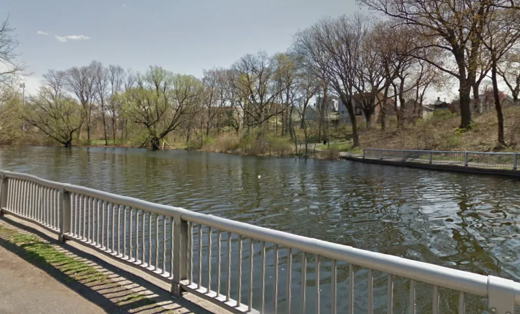 A grey metal railing on the left, next to a path, overlooks a body of water with trees on the far bank and a second railing along that bank.