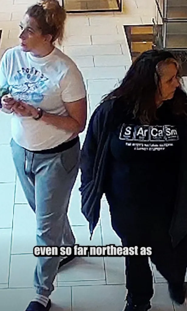 Two women walking, one in a white shirt and gray pants, the other in a black long-sleeved shirt with the word 