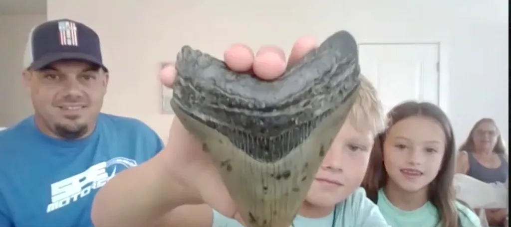 A boy holding up a large fossilized megalodon tooth for the camera, with a man and a girl smiling in the background.