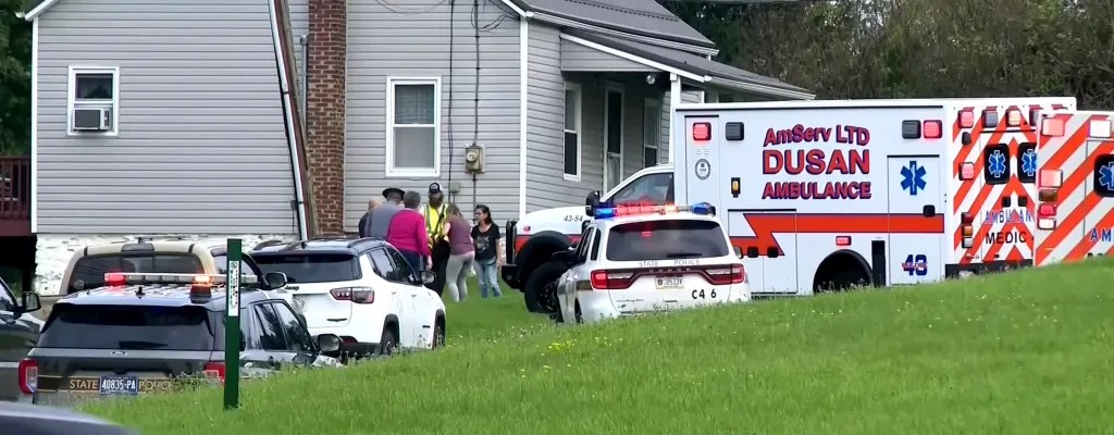 Police cars and an ambulance with flashing lights parked outside a house with people gathered near the entrance.