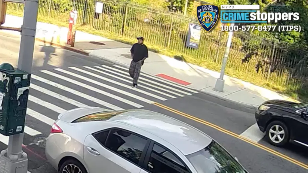Surveillance image of a man in a black shirt and camouflage pants crossing a street, with a white car in the foreground.