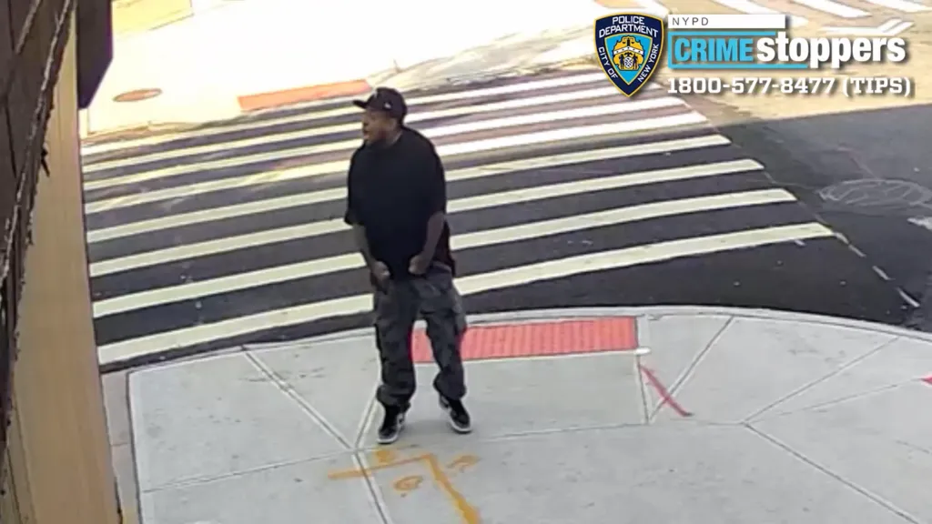 A man in a black shirt, camouflage pants, and a baseball cap stands on a sidewalk.