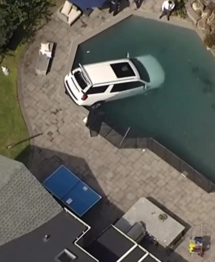 Aerial view of a white SUV submerged in a swimming pool, with the trunk open.