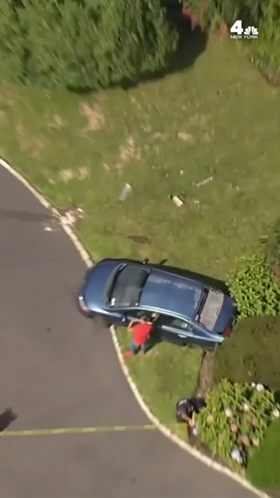 Aerial view of a blue car pulled over on a road shoulder with a man in a red shirt standing by the open driver's side door, and a man in a dark uniform kneeling near a bush.