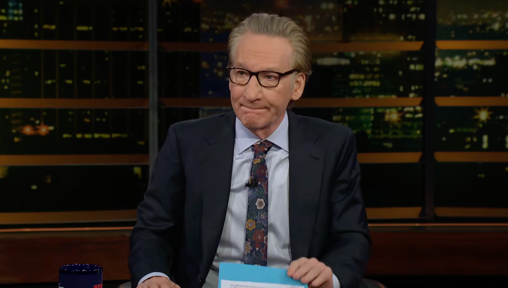 Bill Maher, a white man with glasses and a floral tie, on a talk show set.