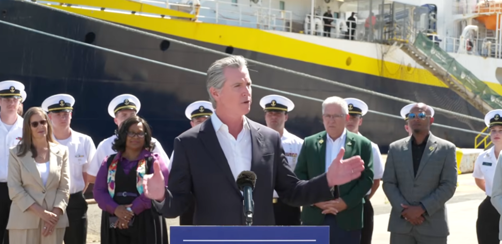 Gavin Newsom speaking at a podium with a ship in the background and officials standing behind him.