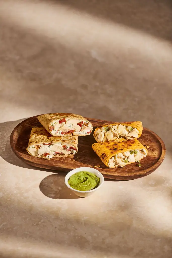 Starbucks Chicken Wrap halves, one with red peppers and one with green chiles, served on a wooden plate with a side of green sauce.