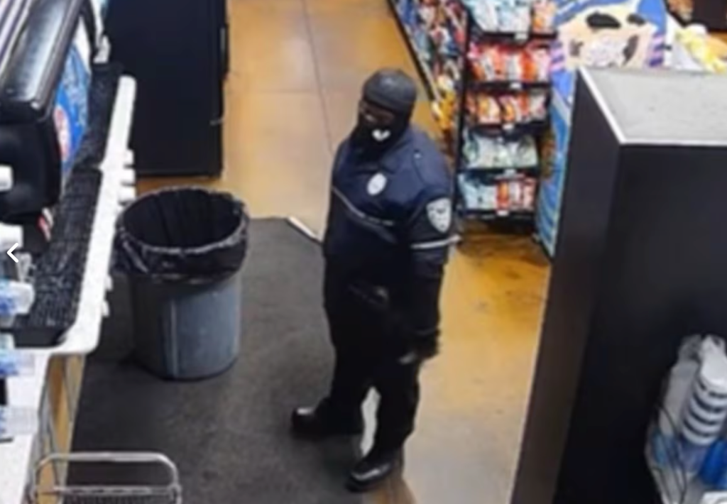 A man in a black and blue uniform, black beanie, and black mask stands in an aisle between a soda machine and snack shelves.