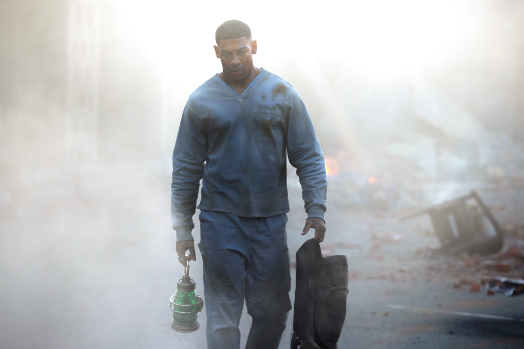 Aaron Pierre in a blue scrub shirt and pants, holding a glowing green lantern and a black bag, walking through a hazy, destroyed landscape.