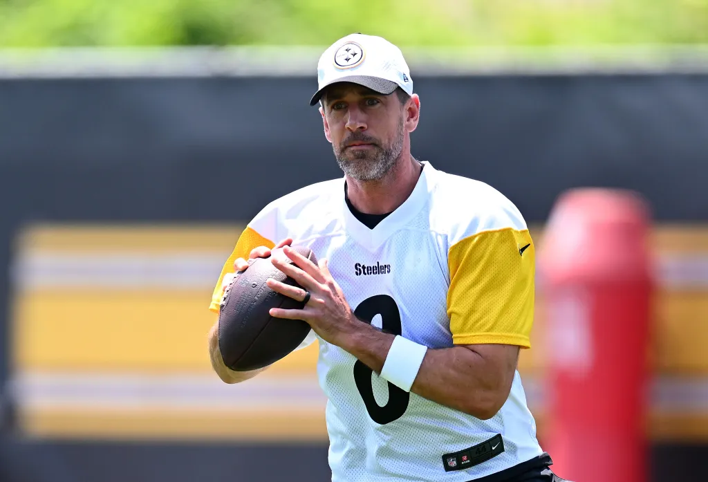 Aaron Rodgers, wearing a white Steelers jersey with yellow sleeves and number 8, holds a football, looking forward.