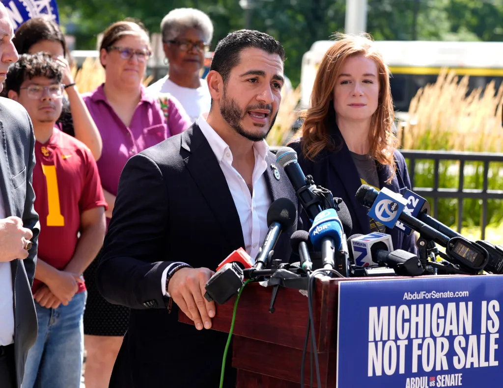 Abdul El-Sayed speaking at a podium with microphones, while Mallory McMorrow listens beside him.