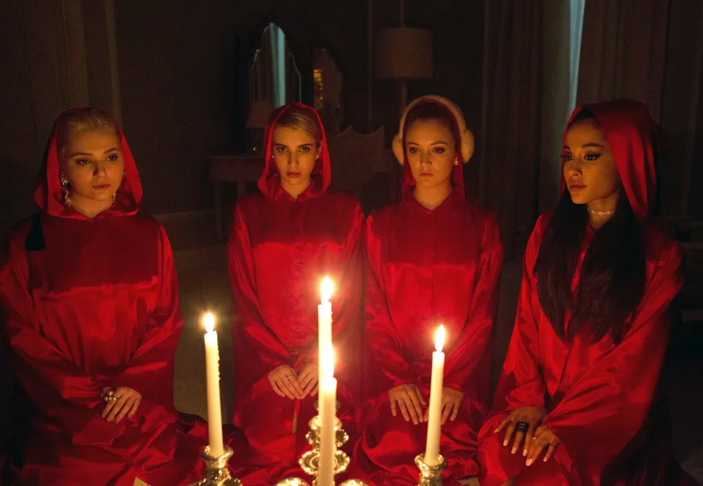 Four women in red robes, Abigail Breslin, Emma Roberts, Billie Lourd, and Ariana Grande, kneel around lit candles.