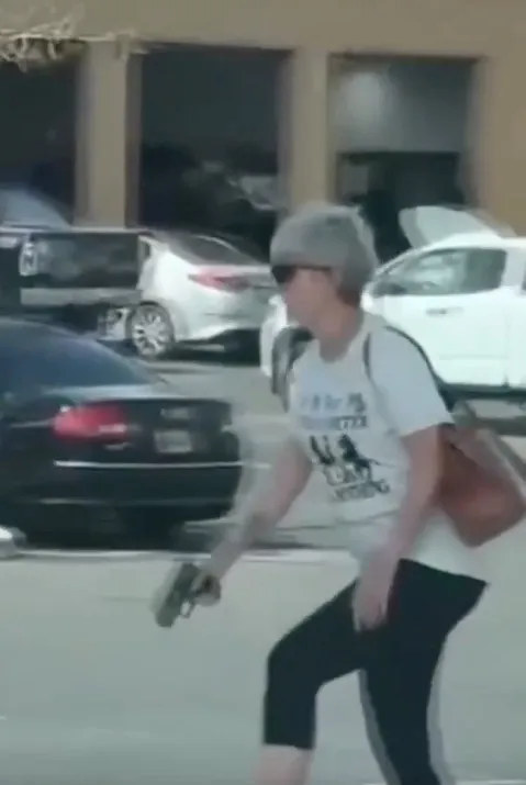 A woman with grey hair and sunglasses, wearing a white t-shirt and dark pants, holds a handgun while walking in a parking lot.