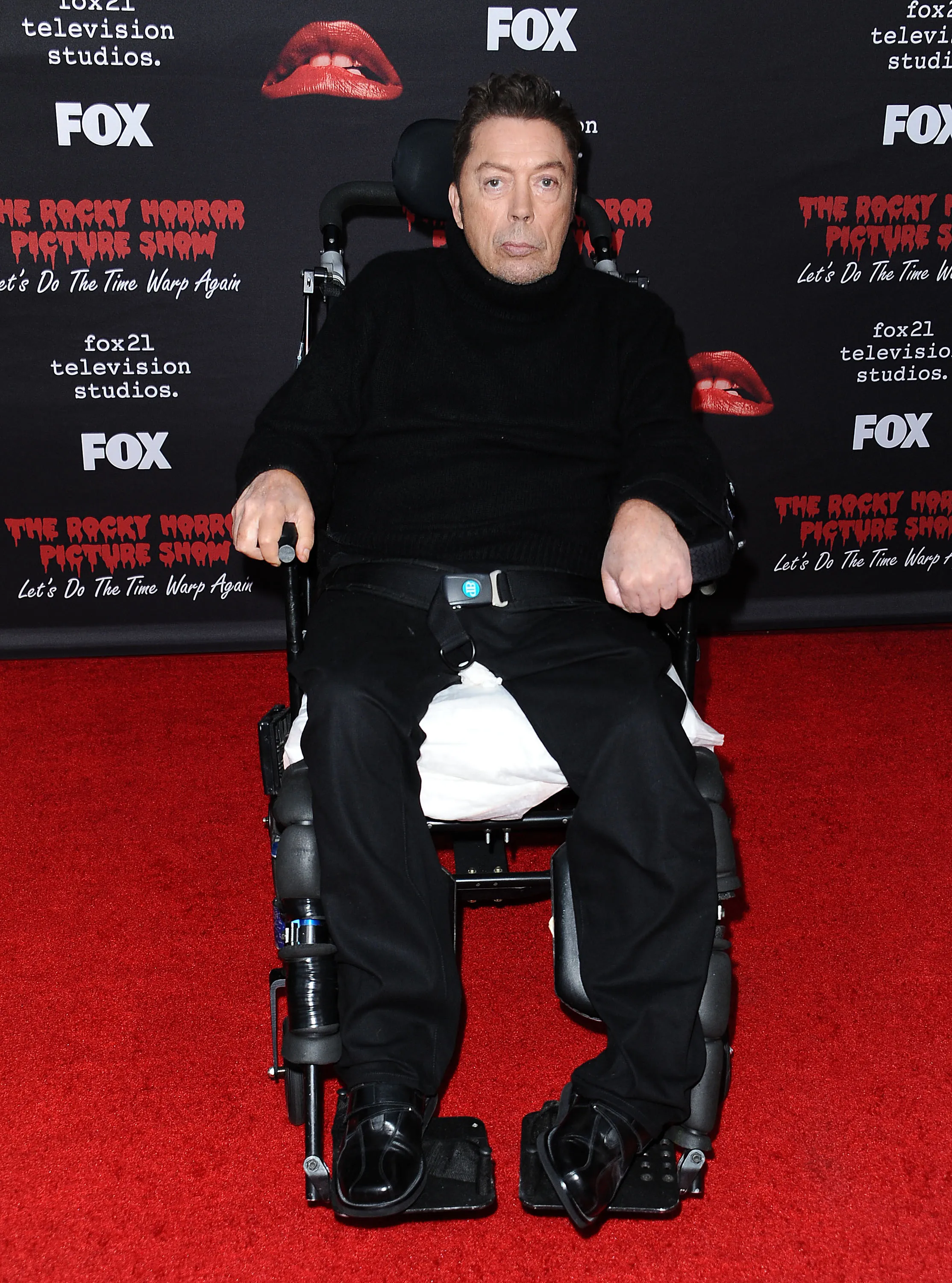 Actor Tim Curry in a wheelchair, wearing a black turtleneck and black pants, at the premiere of