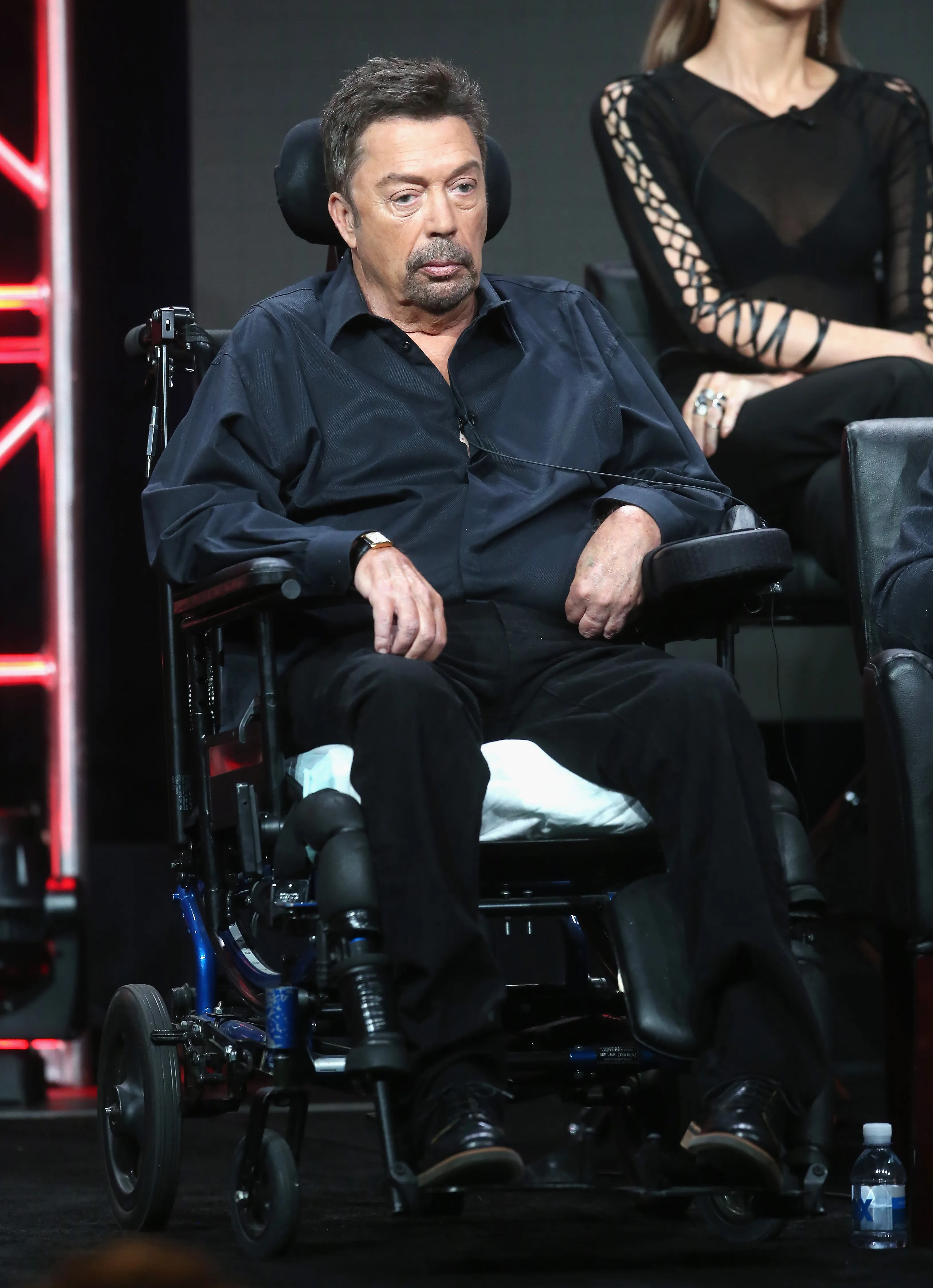 Actor Tim Curry speaks onstage while seated in a wheelchair.