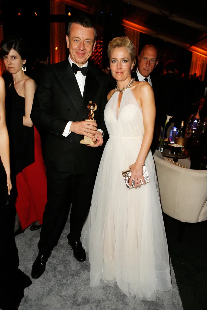 Producer Peter Morgan holding a Golden Globe award, with actress Gillian Anderson in a white dress at a party.
