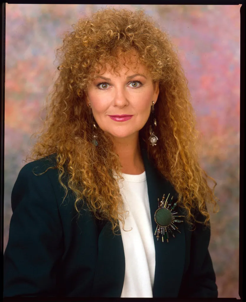 Shelley Fabares smiling, wearing a dark green blazer with a decorative brooch and a white shirt.