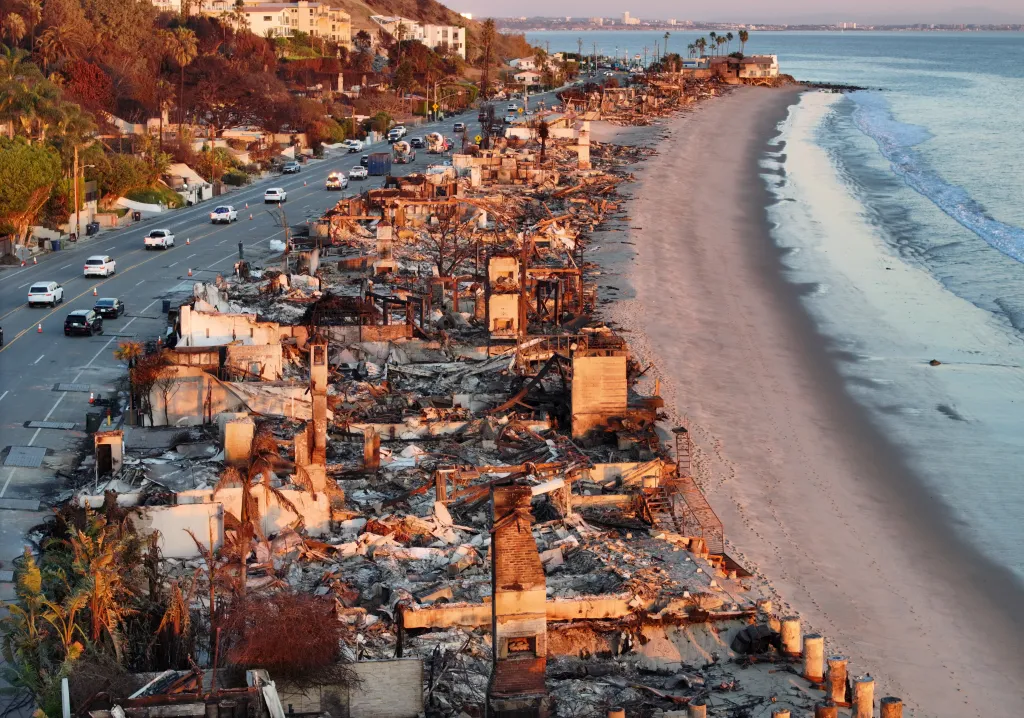 Aerial view of beachfront homes destroyed by wildfire at sunset, with repair vehicles on the road.