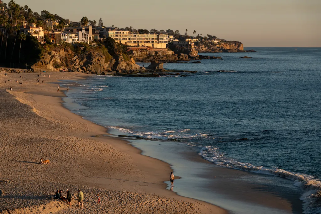 The afternoon sun illuminates Aliso Beach in Laguna Beach, California, with luxury homes perched on cliffs above the sandy shore where people walk and relax near the water.