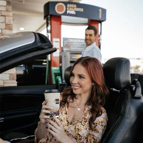 A woman holding a coffee cup in a car with a gas station in the background.