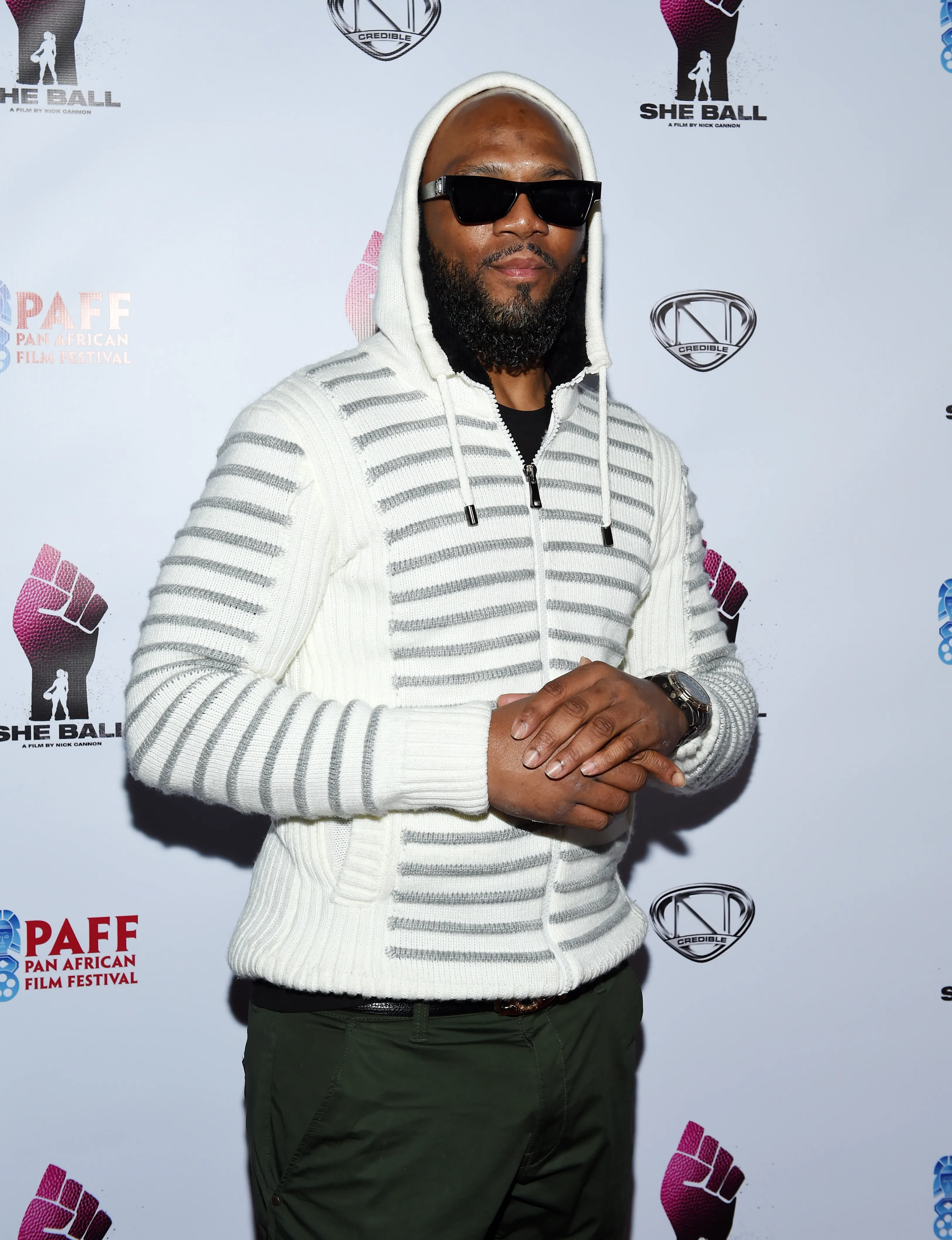 Samuel Monroe Jr. (Caffamilliano) in a white hooded sweater, black t-shirt, dark green pants, and sunglasses.
