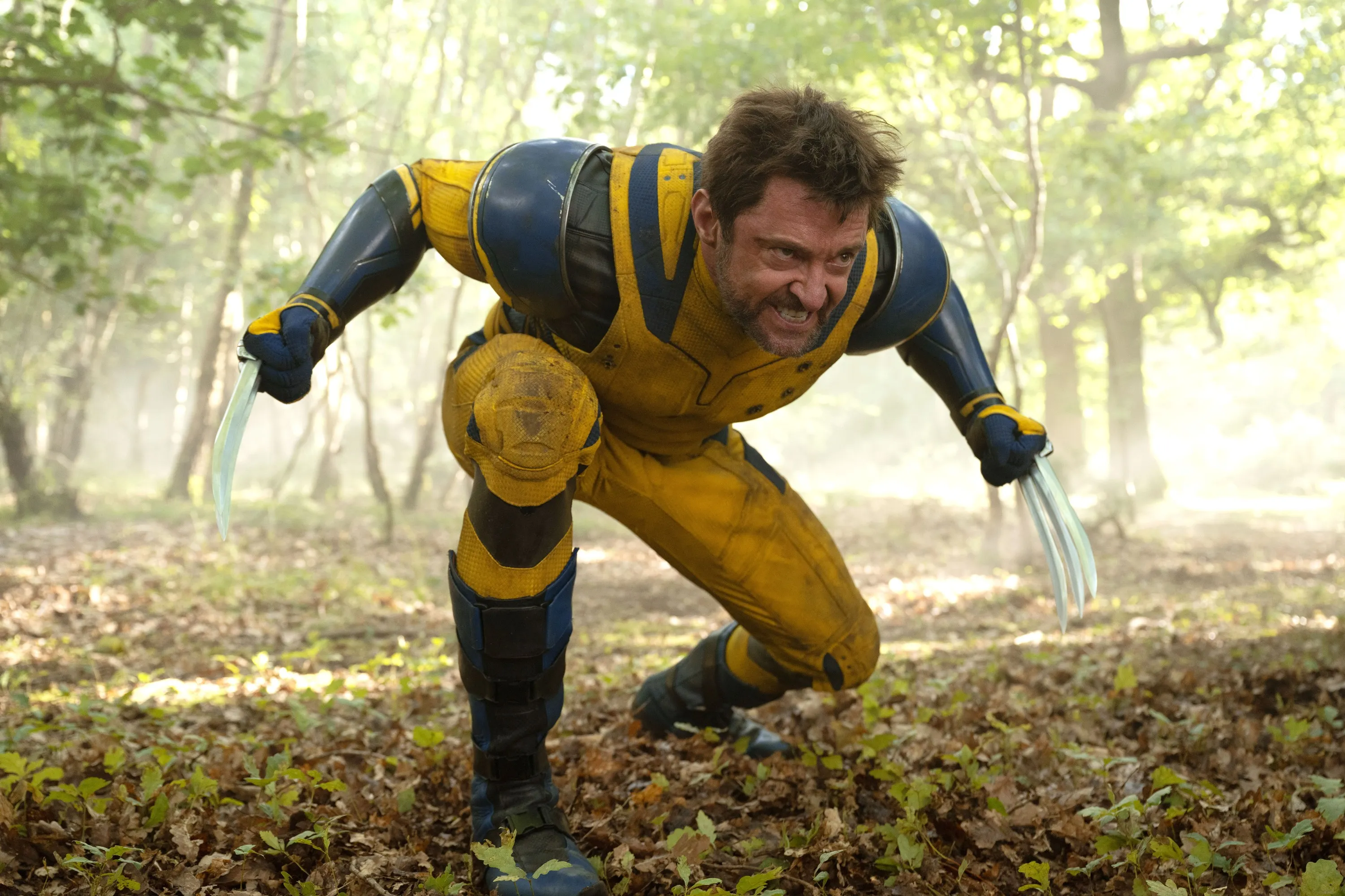 Hugh Jackman as Wolverine in a yellow and blue suit, crouched and grimacing with claws extended, in a forest.