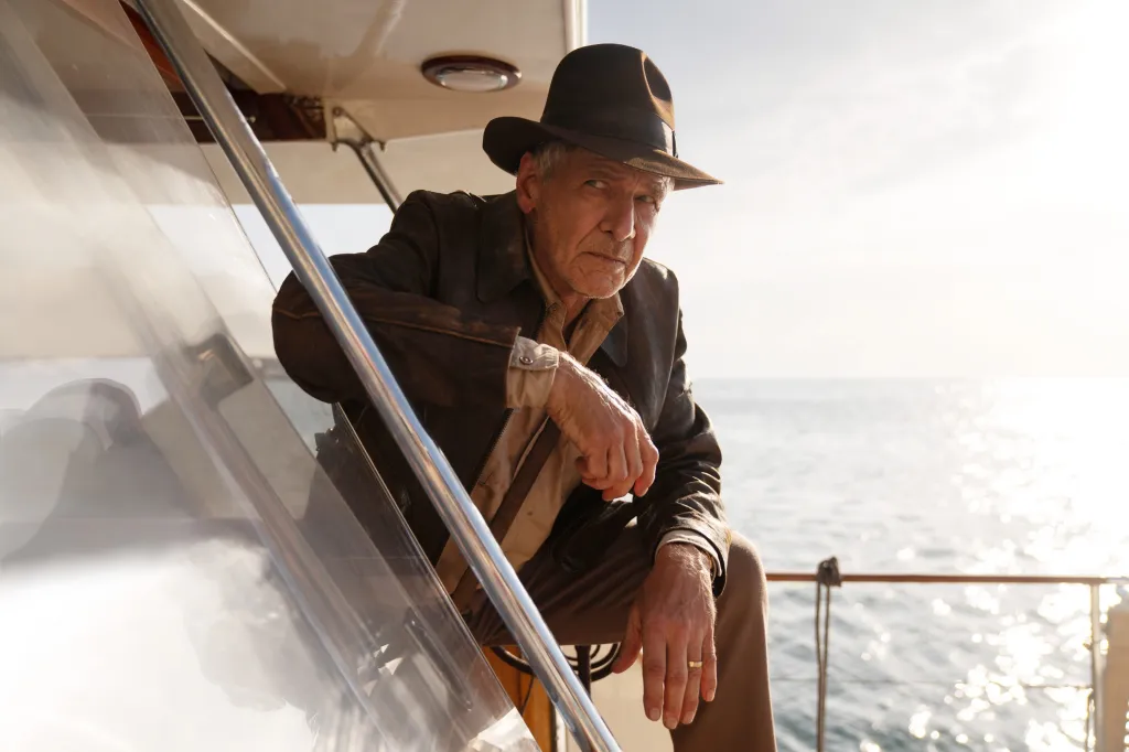 Harrison Ford as Indiana Jones wearing a hat and brown leather jacket, sitting on a boat with the ocean in the background.