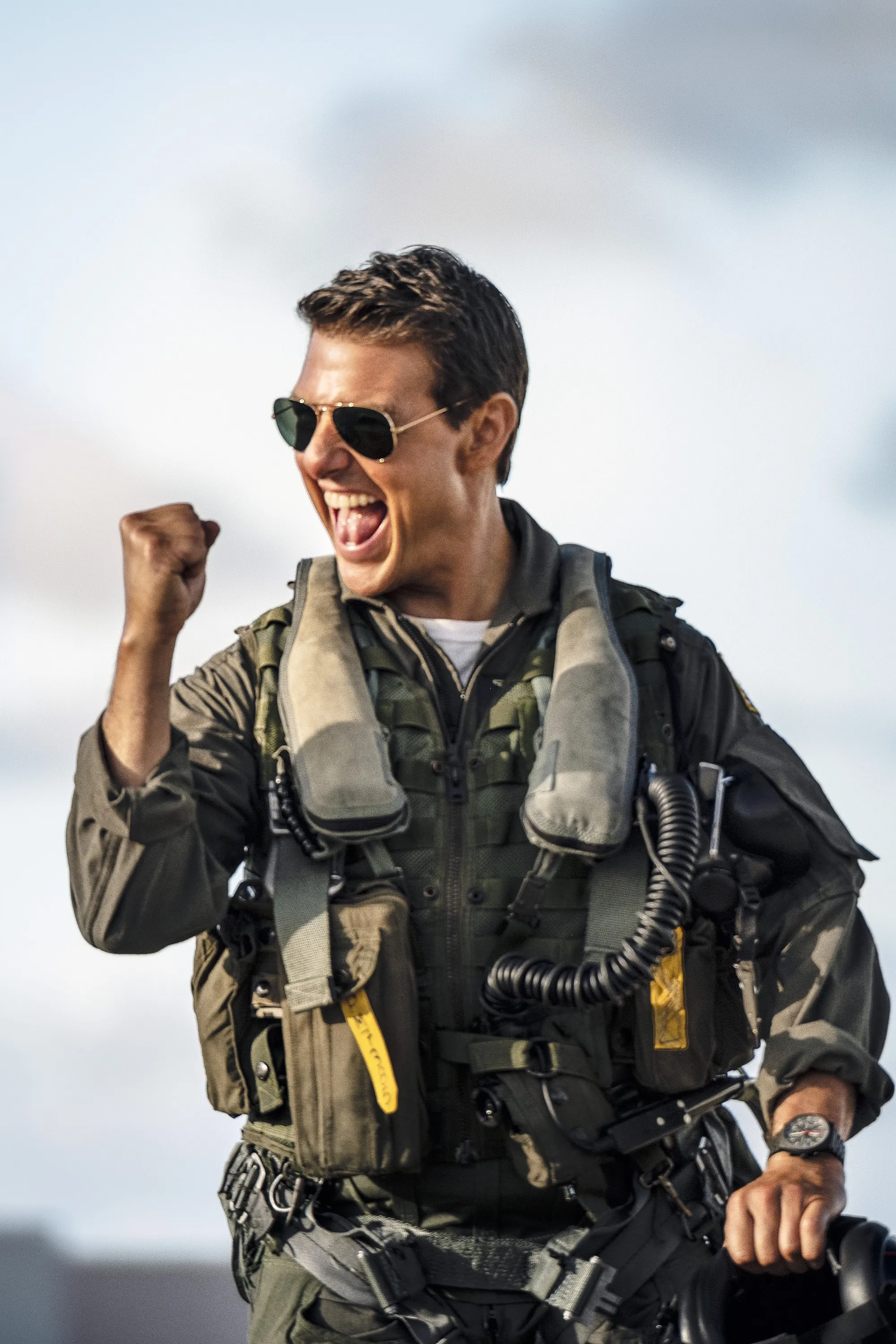 Tom Cruise as Maverick in Top Gun: Maverick, wearing aviator sunglasses and a flight suit, cheering with a clenched fist.