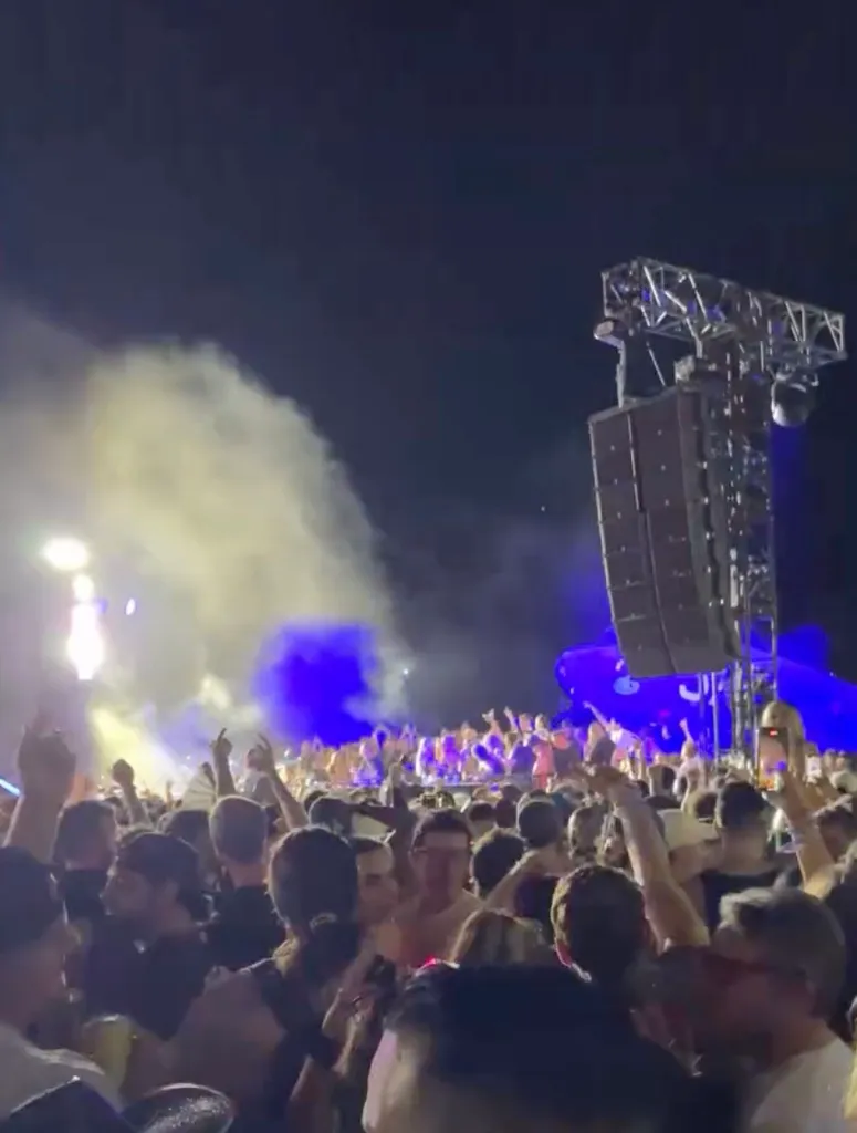 A large crowd of people at an outdoor concert at night with a large speaker tower and smoke.