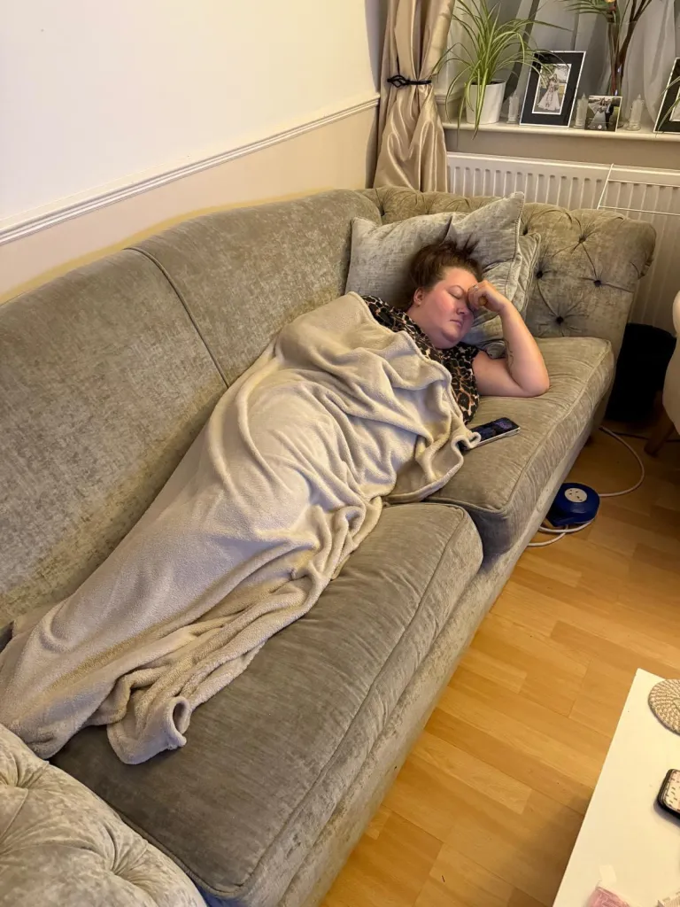 Amber Williams lies on a couch, covered with a blanket, holding her head in pain.