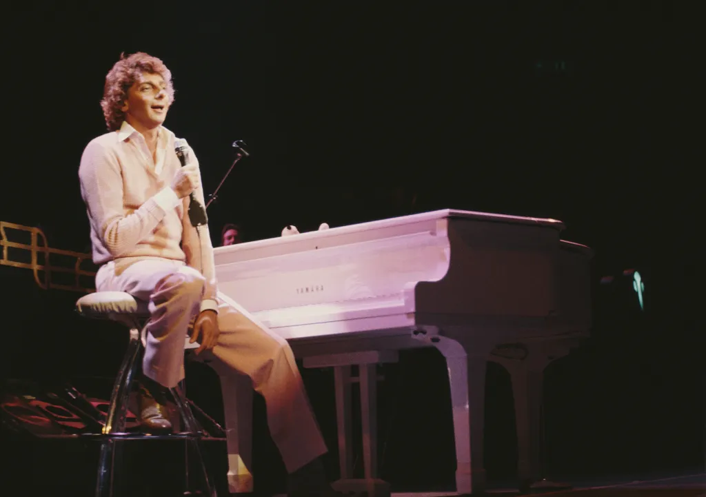 Singer-songwriter Barry Manilow performing live in concert while seated on a stool with a microphone and white piano.