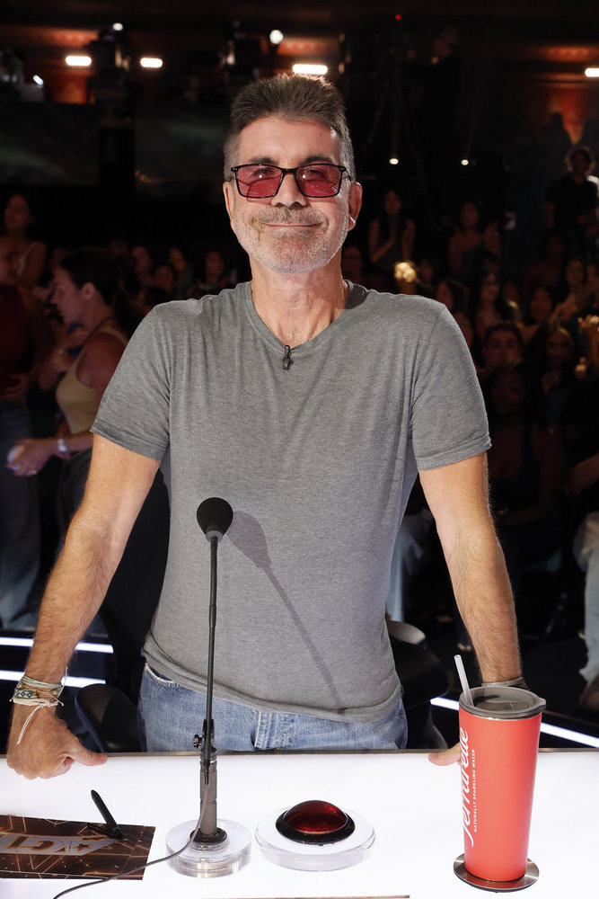 Simon Cowell from America's Got Talent, wearing a gray t-shirt and pink-tinted glasses, stands behind a judge's desk with a red buzzer and a red cup.