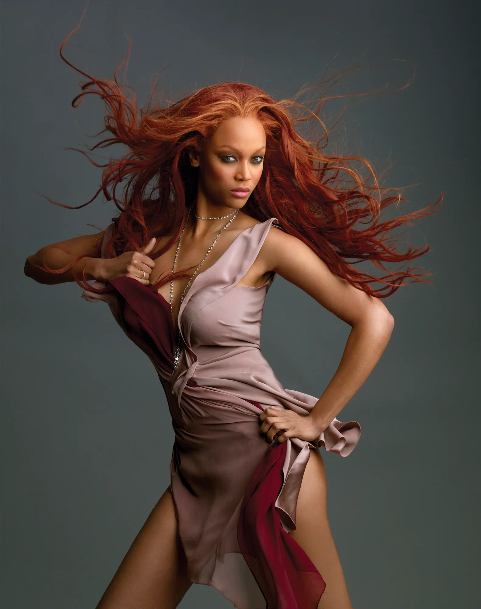 Tyra Banks with long, flowing reddish-brown hair, wearing a beige and dark red dress, posing against a dark background.