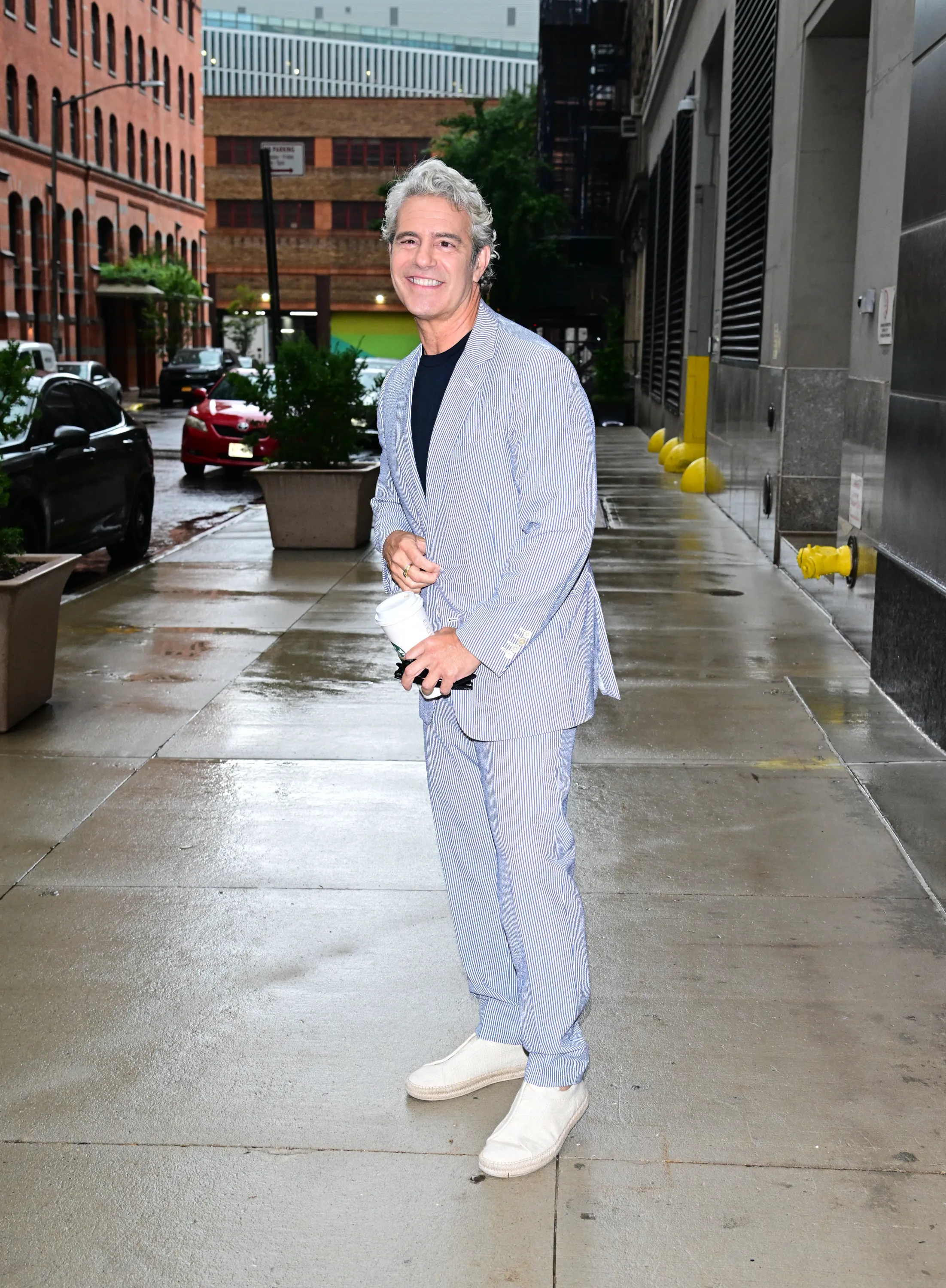 Andy Cohen standing on a wet sidewalk in a blue and white pinstriped suit.