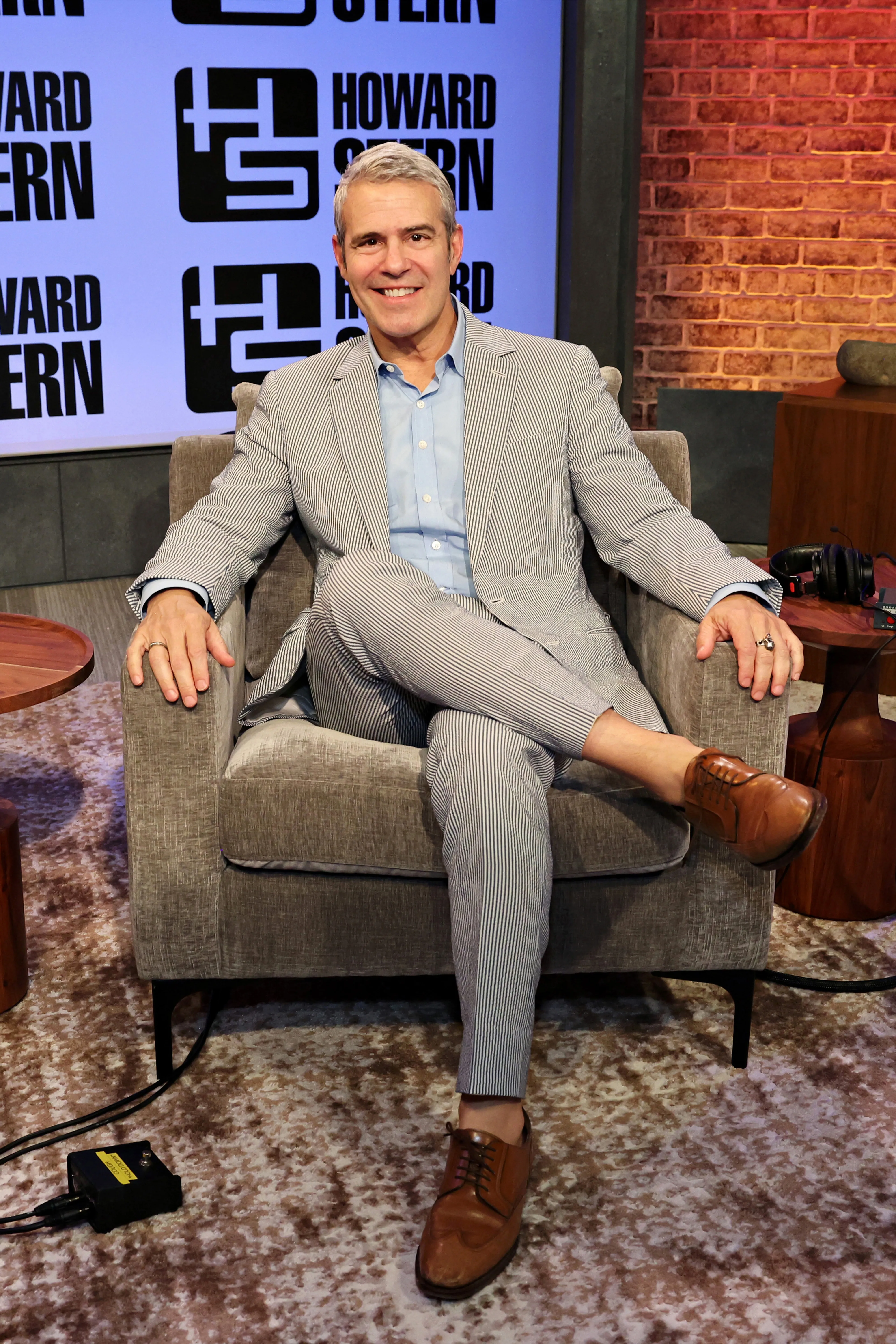 Andy Cohen in a striped suit and light blue shirt, seated in a grey armchair, smiling.