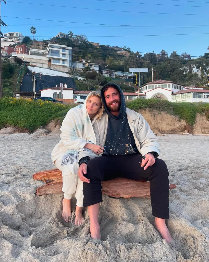 Annalynne Mccord and Danny Cipriani sitting together on a log on a sandy beach.