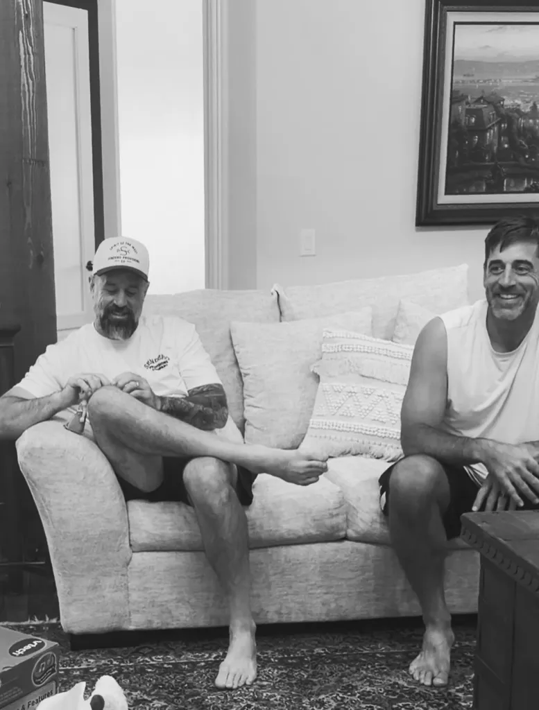 Aaron Rodgers sits on a sofa next to another man, smiling while the man on the left looks at his feet.