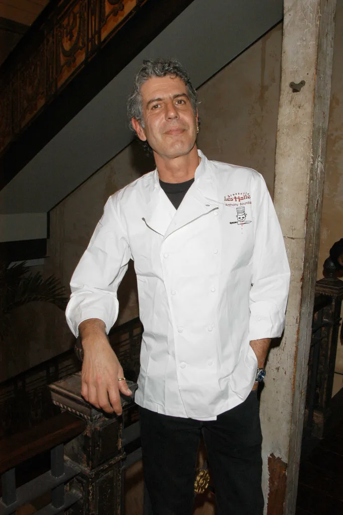 Anthony Bourdain in a white chef's coat, leaning on a dark wooden railing.