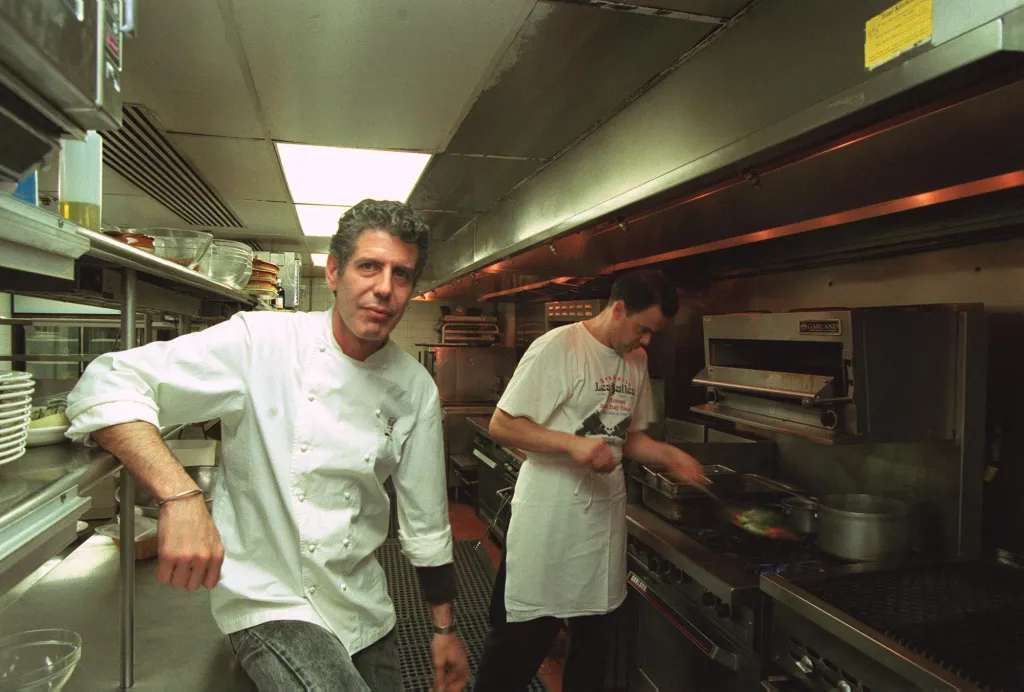 Anthony Bourdain in a white chef's coat leans against a counter in a professional kitchen, looking at the camera, while another chef cooks in the background.