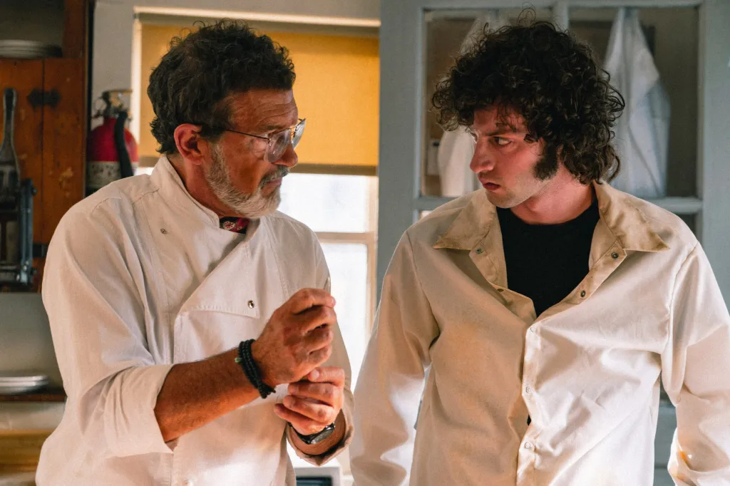 Antonio Banderas as a chef, with a beard and glasses, looking at Dominic Sessa as Anthony Bourdain with curly hair, in the movie 