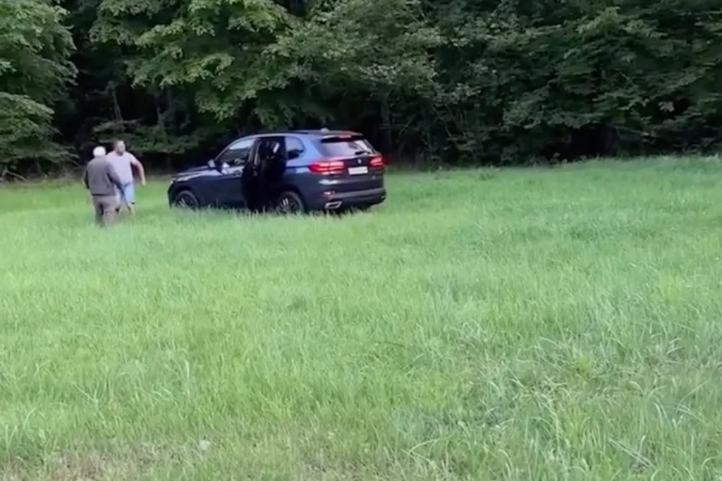 A man in a white shirt and a man in a gray shirt stand in a grassy field next to an SUV with its door open.