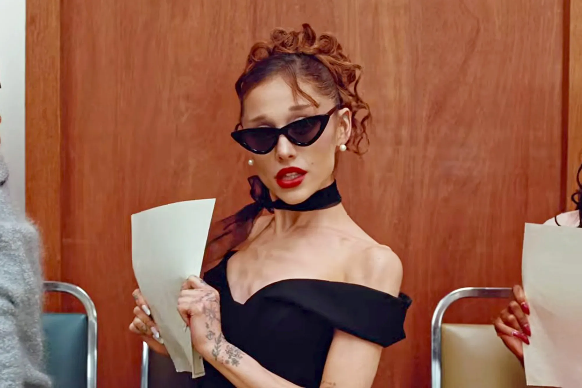 Ariana Grande in a black off-the-shoulder top, black cat-eye sunglasses, and a black choker, holding a sheet of paper.