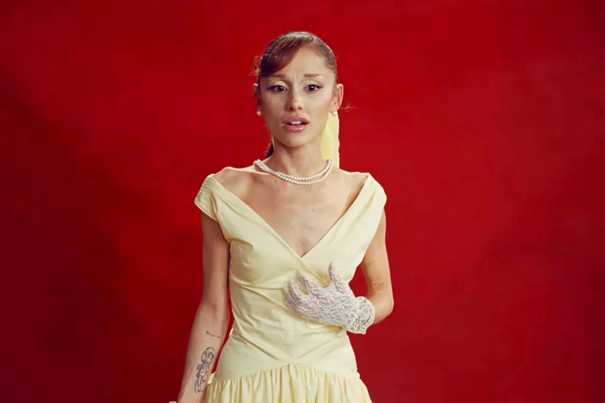 Ariana Grande in a yellow dress, pearl necklace, and white lace gloves against a red background.