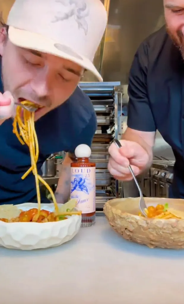 Brooklyn Beckham eating spaghetti from a white bowl next to a man eating from a wooden bowl.