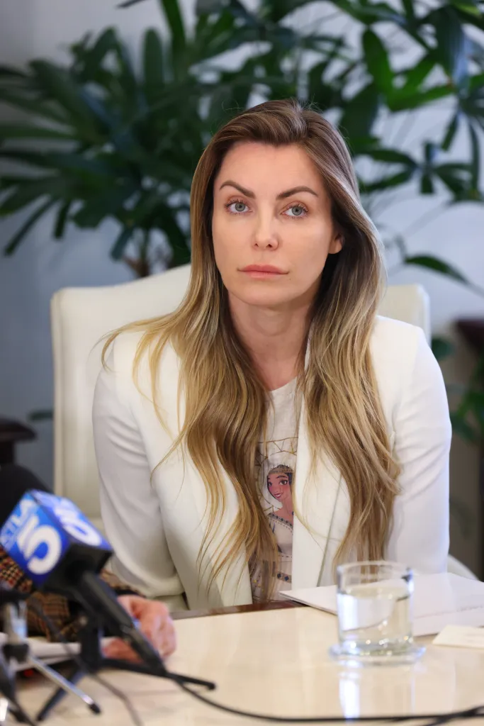 Crystal Hefner looks directly at the camera while sitting at a table with microphones in front of her.